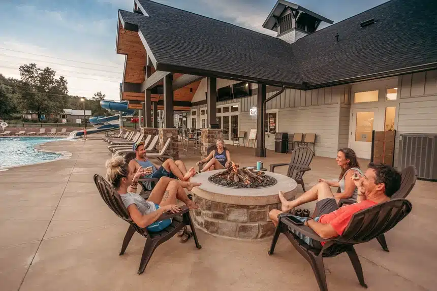 Guests relaxing around an outdoor fire pit beside a resort-style RV campground pool and clubhouse