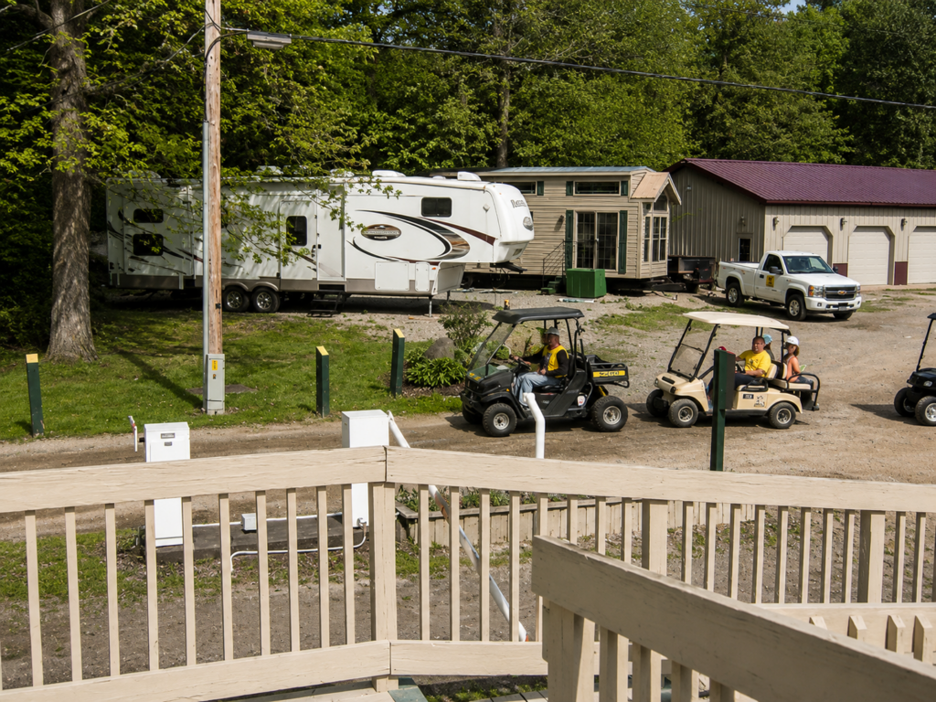 RV campers driving golf carts through a wooded campground with a fifth-wheel RV, cabin-style lodging, and campground facilities on a sunny afternoon.