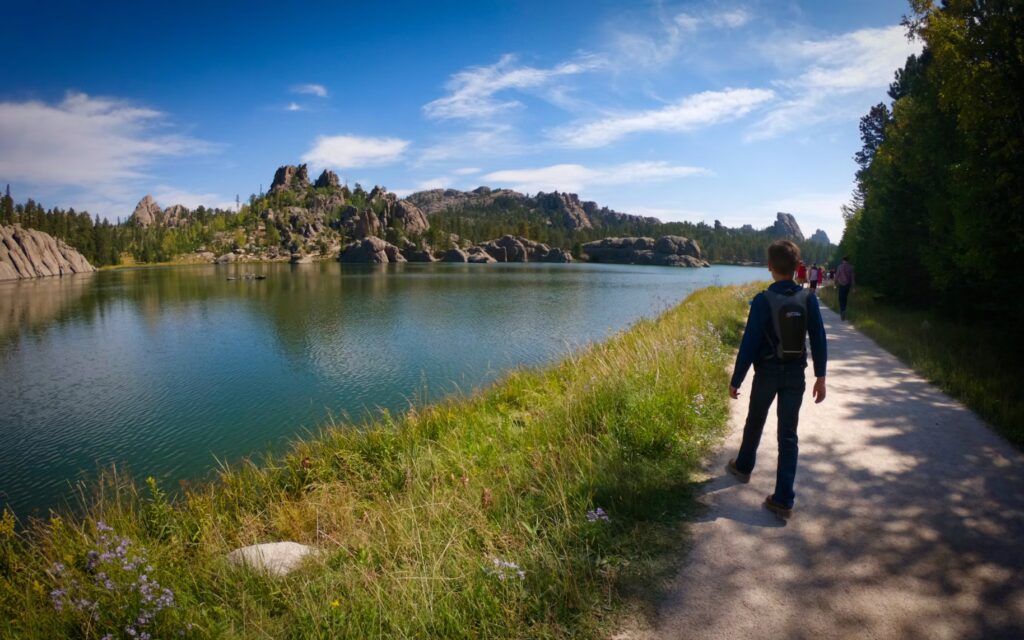 Person walking along lakeside trail with rock formations and clear sky