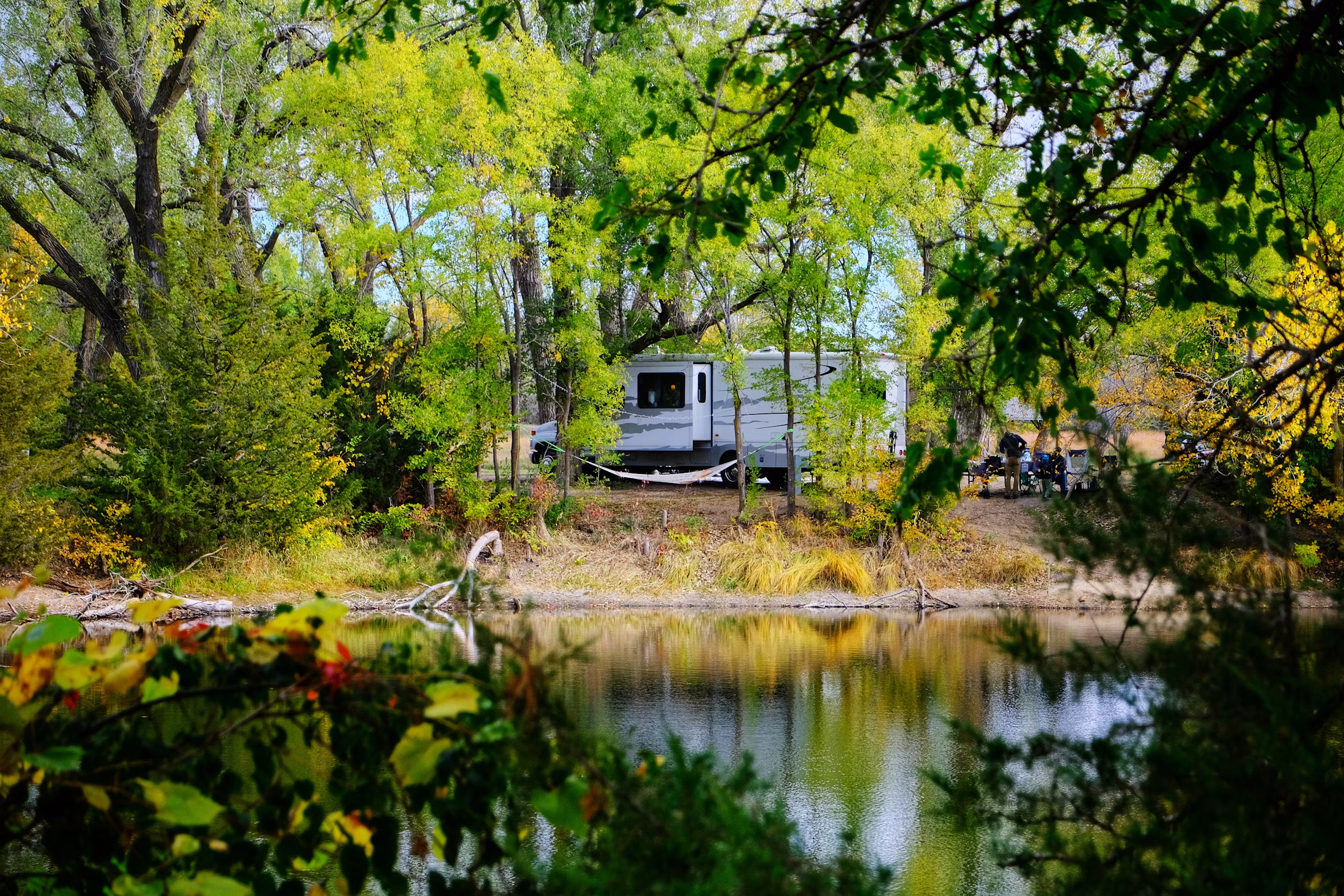 RV camper parked beside a quiet pond surrounded by lush green trees and early fall foliage, reflected in calm water at a wooded campground.