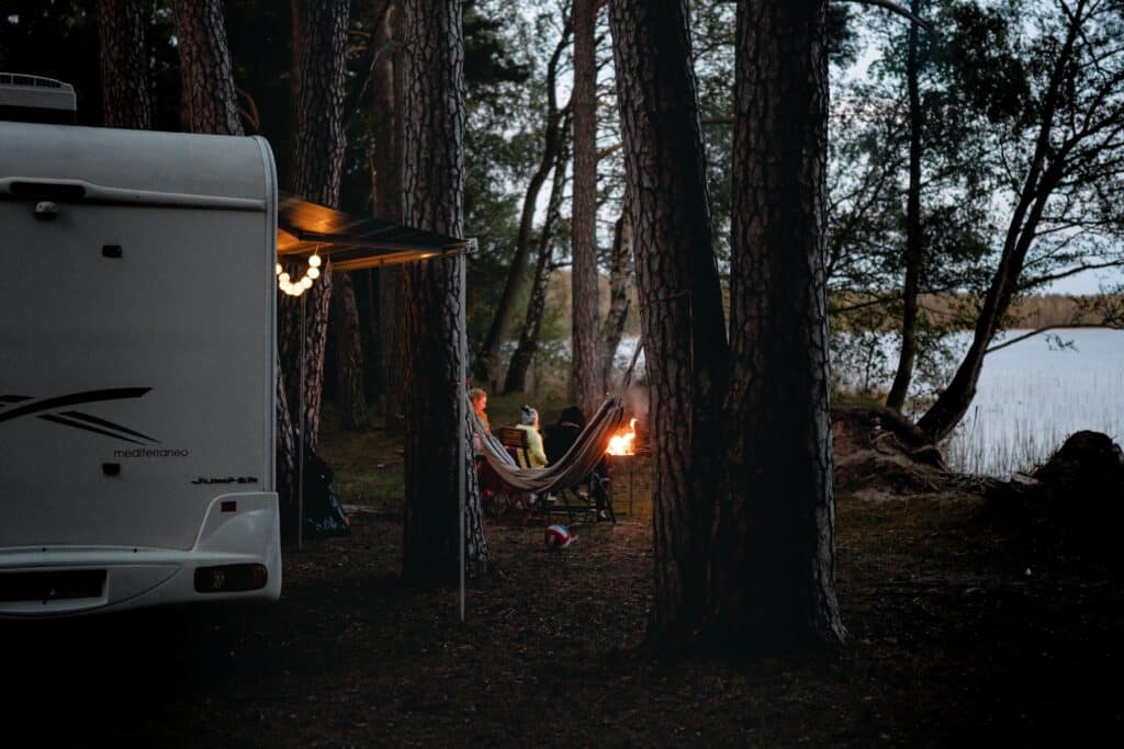 RV campsite set up beside a peaceful lake with string lights, a hammock, and a campfire surrounded by tall pine trees at dusk.
