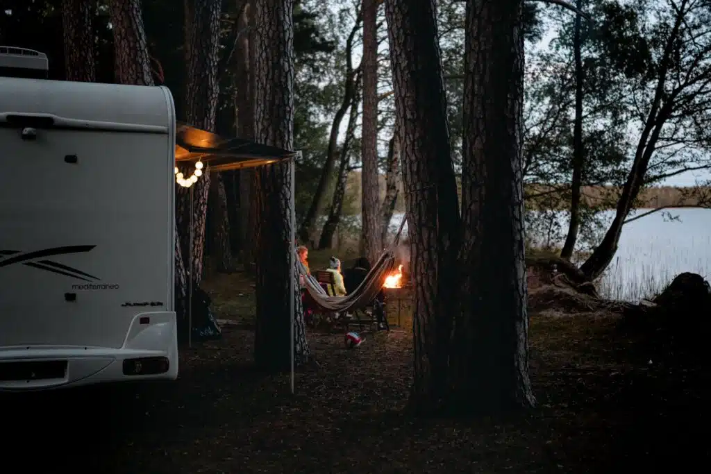 RV campsite set up beside a peaceful lake with string lights, a hammock, and a campfire surrounded by tall pine trees at dusk.